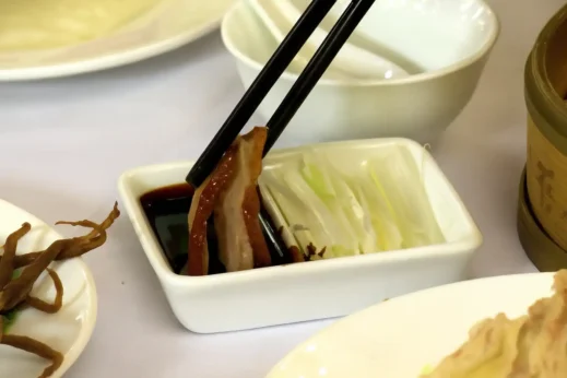 Close-up of Peking duck being dipped in dark sauce with chopsticks beside sliced scallions in a white dish, surrounded by traditional Chinese tableware on a white tablecloth.